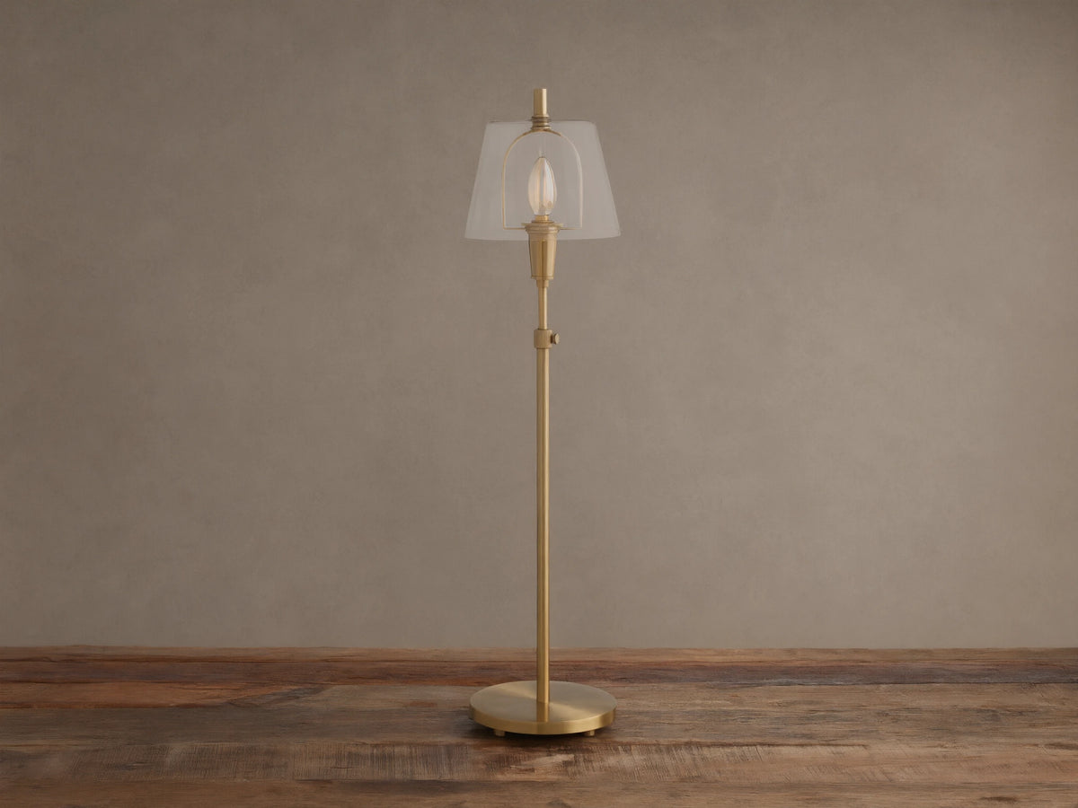 Brass Candice Table Lamp with a clear conical shade and illuminated bulb, on a wooden surface against a textured beige wall.
