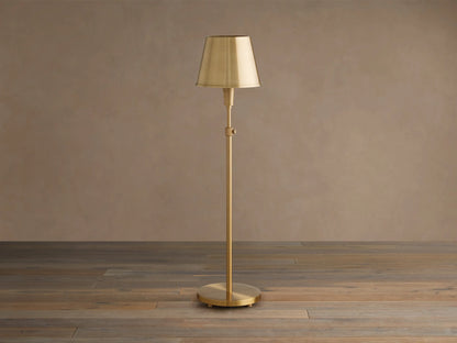Illuminated Candice Table Lamp in brass, with a cone-shaped brass shade, standing on a wooden floor against a beige wall.