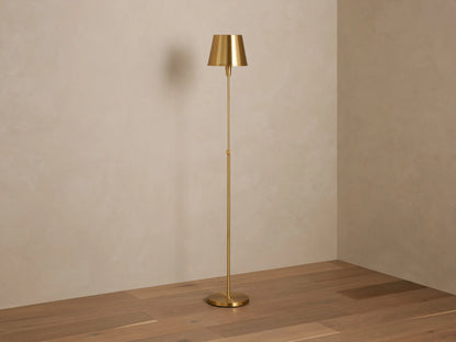 Candice Floor Lamp in brass finish with a tapered shade, lit and casting a warm glow, standing in a room corner on a wooden floor.