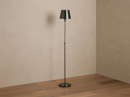 Candice Floor Lamp with a slender dark pewter-finished stand & tapered shade, illuminated, in a room corner with wood floor.