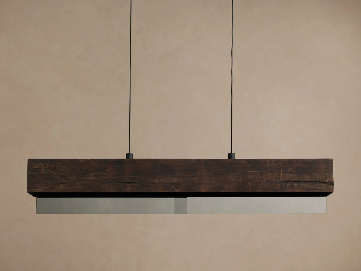 Lena Linear Chandelier: A rectangular dark wood beam with textured grain, featuring mirrored glass below, emitting a warm glow, suspended by black cords.