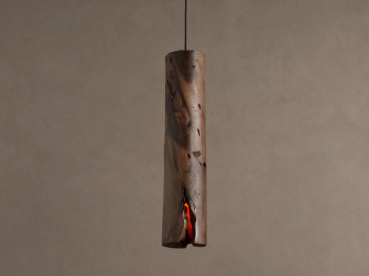 Dedalo Pendant, a cylindrical milpa wood light with natural texture, hangs vertically. An orange glow emanates from a front crack.