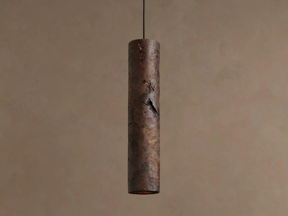 Dedalo Pendant, a cylindrical milpa wood light with natural texture, hangs vertically. An orange glow emanates from a front crack.