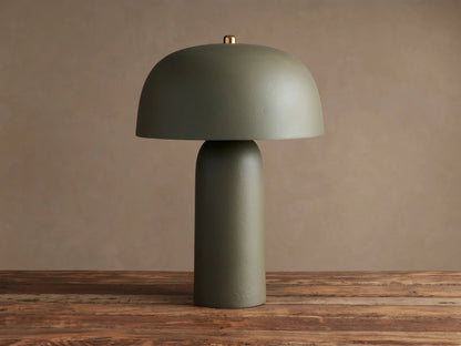 Ira Table Lamp: Sage green lamp with a dome shade, cylindrical base, and small brass accent, emitting a warm glow on wood.
