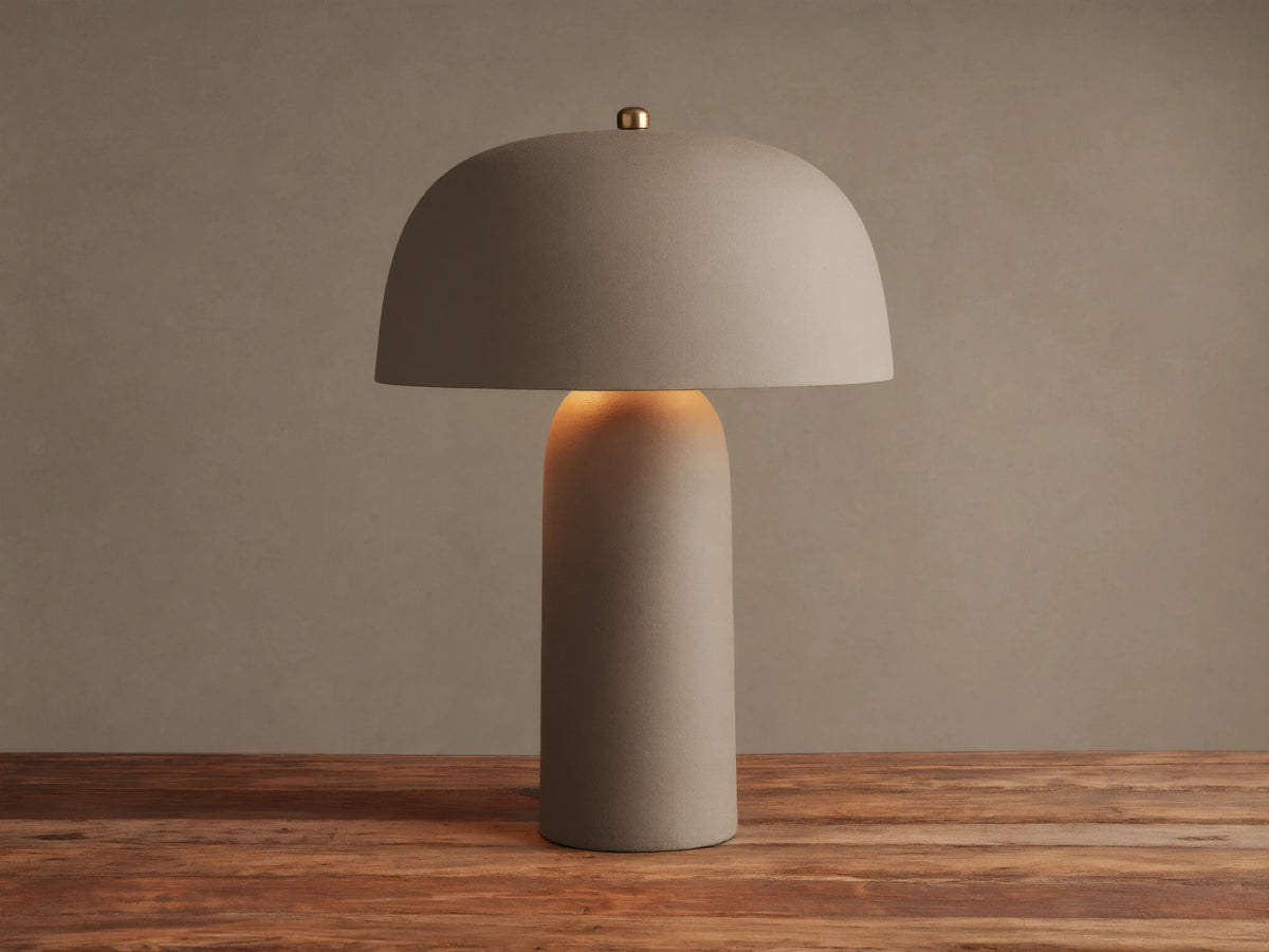 Ira Table Lamp, a textured taupe mushroom-shaped lamp with a gold accent, casting a warm glow on a wooden table.
