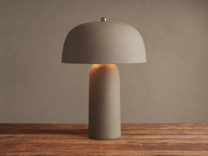 Ira Table Lamp, a textured taupe mushroom-shaped lamp with a gold accent, casting a warm glow on a wooden table.