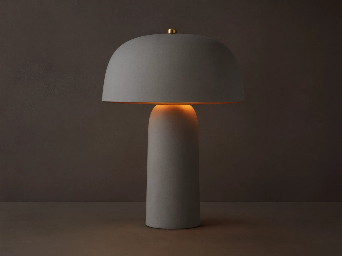 Ira Table Lamp, a textured taupe mushroom-shaped lamp with a gold accent, casting a warm glow on a wooden table.