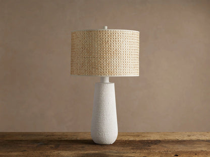 Stucco Table Lamp with a textured white conical base and a natural woven cane cylindrical shade, on a rustic wooden table.