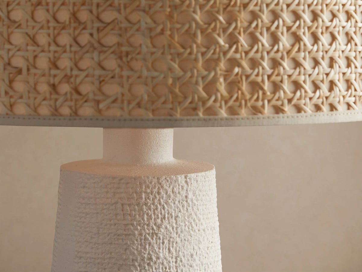 White stucco table lamp with a textured tapered white base and cylindrical natural woven cane shade, on a rustic wood surface.
