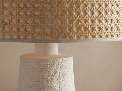 White stucco table lamp with a textured tapered white base and cylindrical natural woven cane shade, on a rustic wood surface.