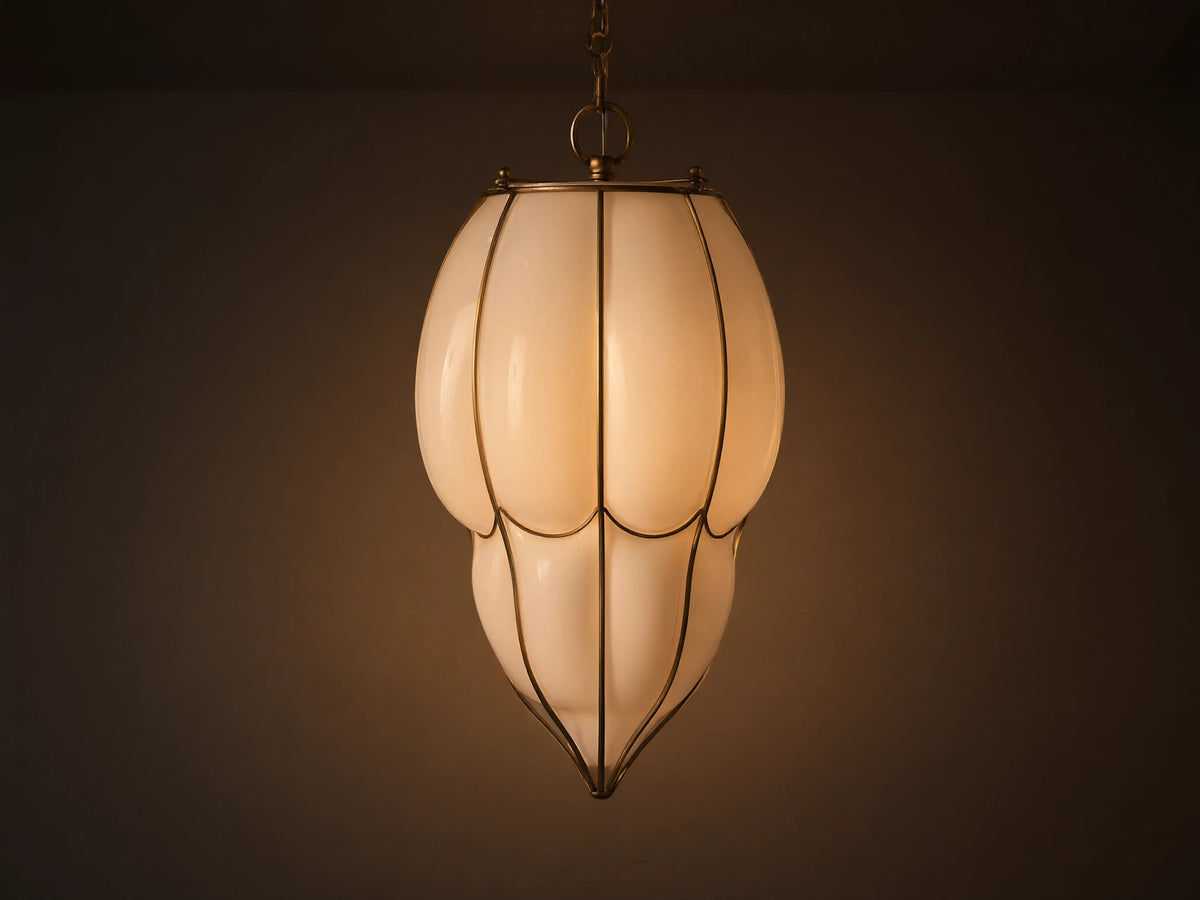 Margolis Large Pendant: Elongated white opaque glass pendant light with brass-finished frame, glowing softly, against a neutral wall.