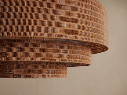 Paloma Chandelier, tiered warm brown handwoven rattan shade hanging from a dark metal chain against a plain beige wall.