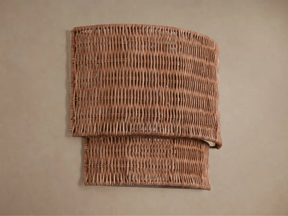 Paloma Sconce with a curved, layered handwoven rattan shade in natural brown, glowing warmly on a beige wall.