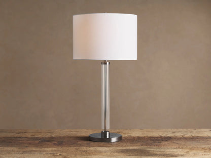Glass Column Table Lamp with clear glass column, brushed metal base, and white linen shade on a wooden table.