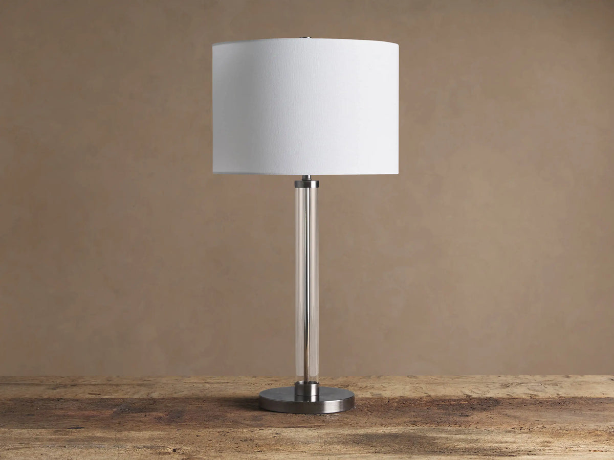 Glass Column Table Lamp with clear glass column, brushed metal base, and white linen shade on a wooden table.