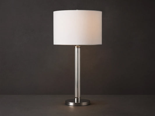 Glass Column Table Lamp with clear glass column, brushed metal base, and white linen shade on a wooden table.