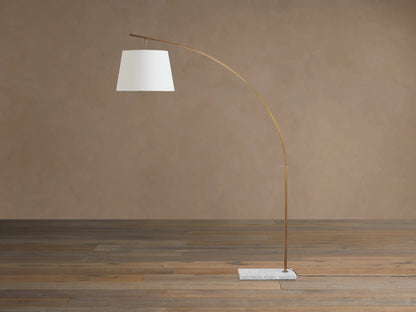 Tadeo arc floor lamp features a curved brass-colored stand, white linen drum shade, and square white marble base on a wood floor.