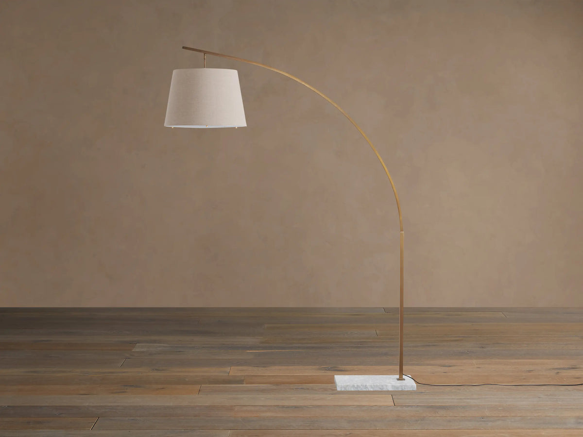 Tadeo Floor Lamp with an arched brass arm, cream linen drum shade, and white marble base on a wooden floor.