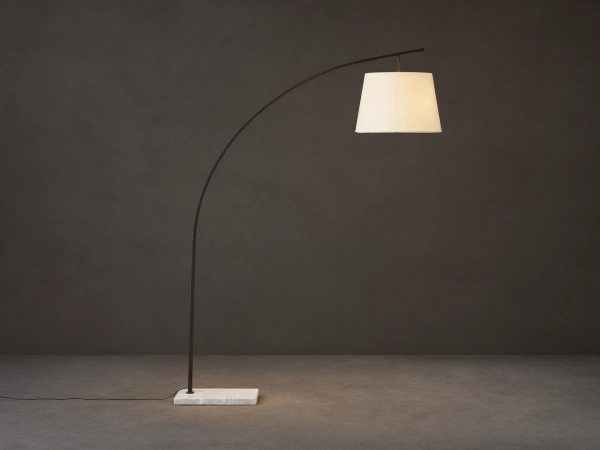 Tadeo Floor Lamp with a curved dark bronze arm, white linen shade, and rectangular white marble base on a wood floor.