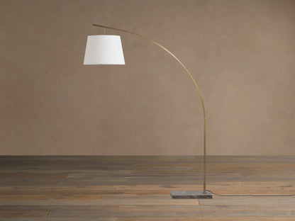 Tadeo Floor Lamp with arcing brass-toned pole, white linen shade, and dark marble base on a wooden floor.
