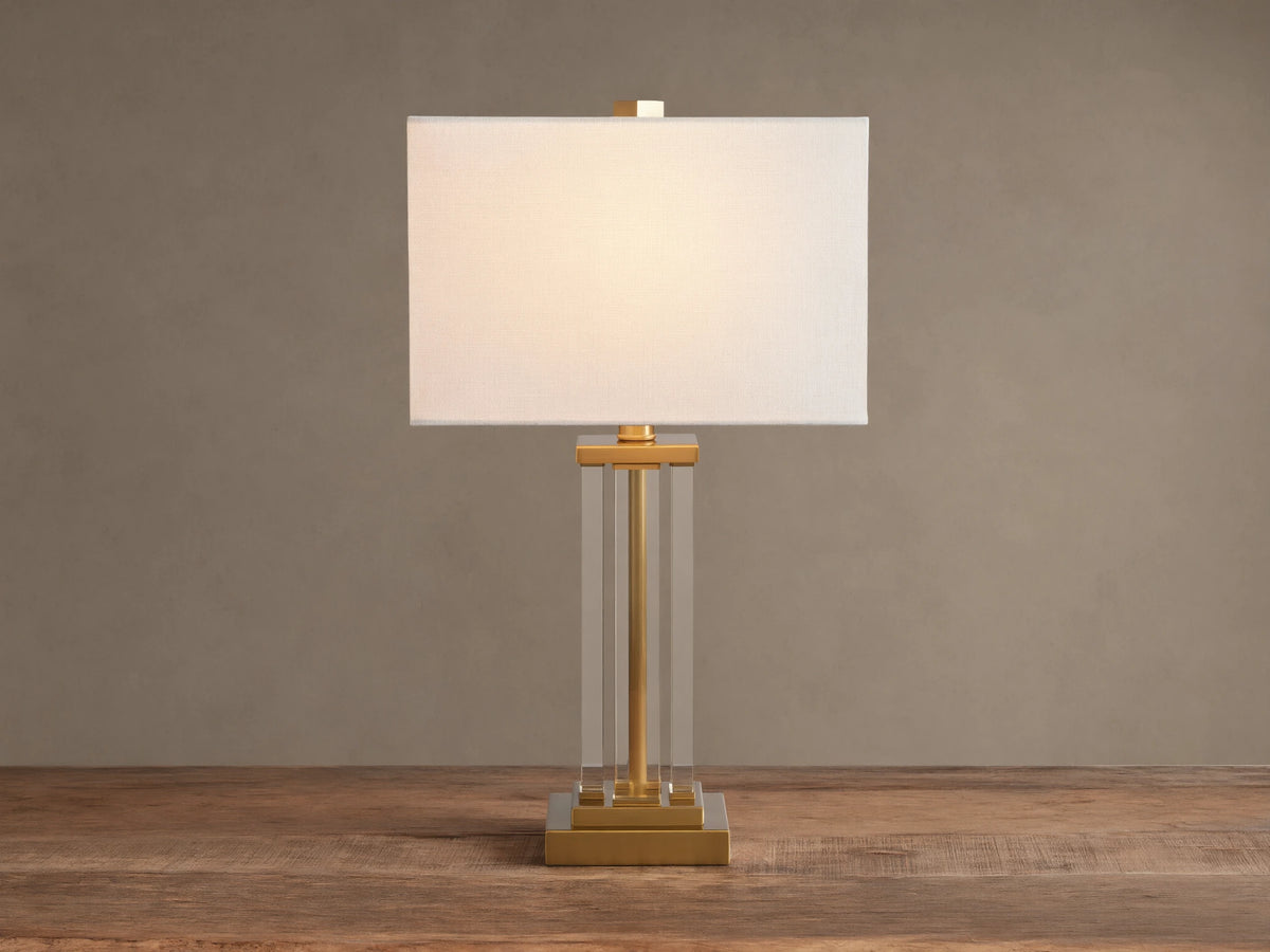 Dawson Table Lamp: white rectangular shade, clear acrylic column body, and gold-tone square base on a wood surface.