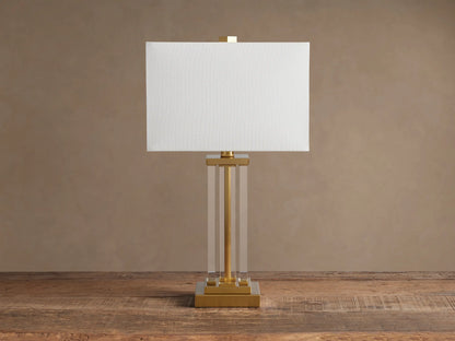 Dawson Table Lamp: white rectangular shade, clear acrylic column body, and gold-tone square base on a wood surface.