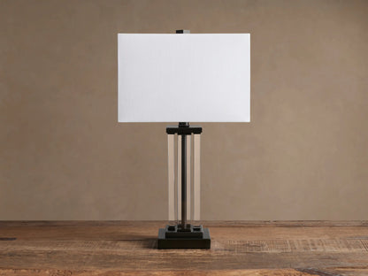 Dawson Table Lamp with a lit rectangular white fabric shade, clear acrylic pillars, and a dark bronze square base on a wooden table.
