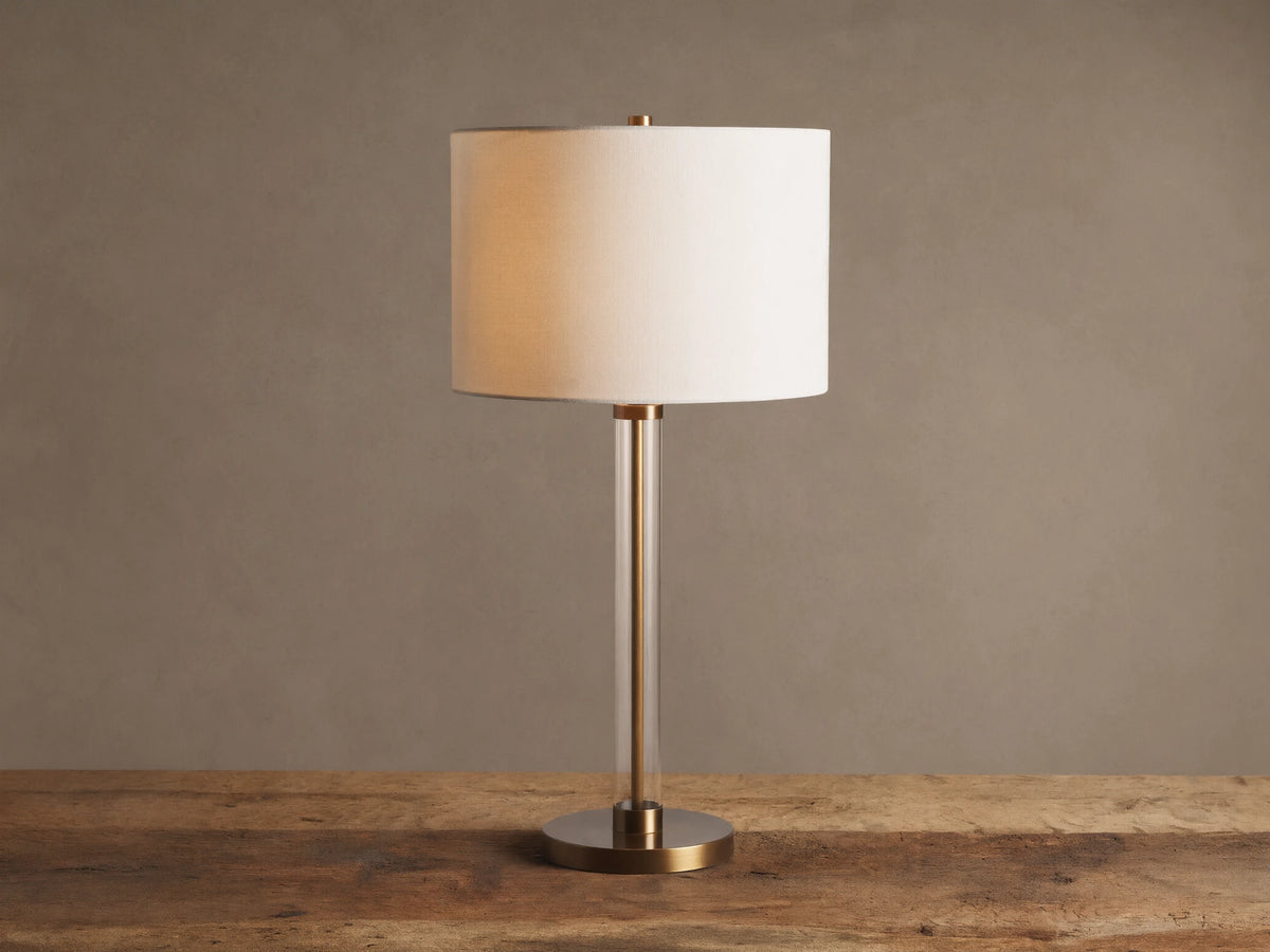 Lit Glass Column Table Lamp featuring a clear glass stem, bronze base, and cream linen drum shade on a rustic wood table.