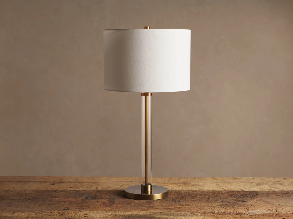 Lit Glass Column Table Lamp featuring a clear glass stem, bronze base, and cream linen drum shade on a rustic wood table.