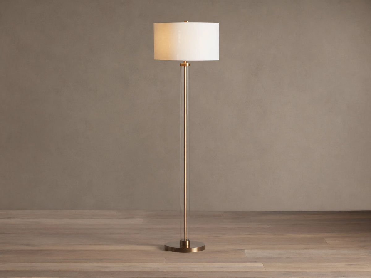 Clear glass column floor lamp with a brass base and stem, and a cylindrical cream linen shade. Standing on a wooden floor.