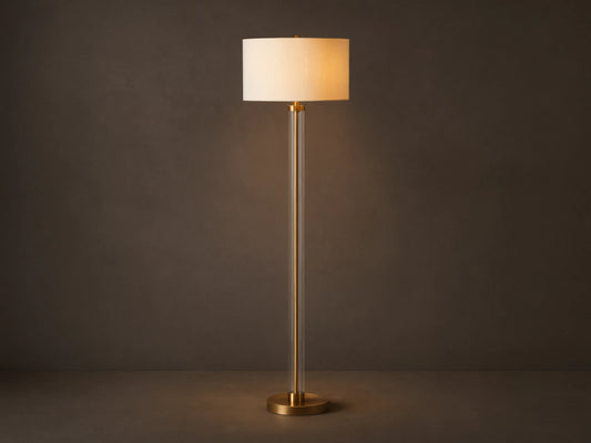 Clear glass column floor lamp with a brass base and stem, and a cylindrical cream linen shade. Standing on a wooden floor.