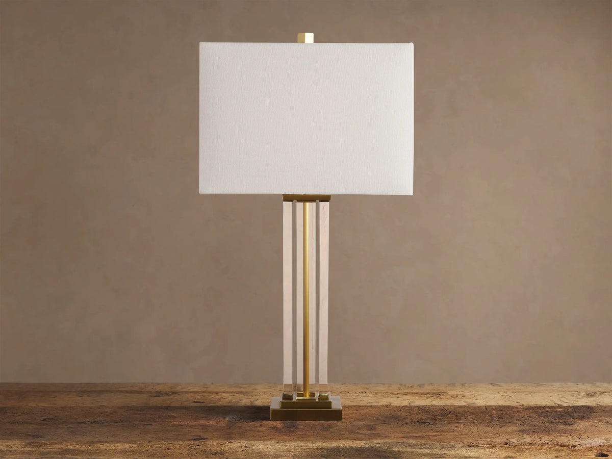 Dawson Table Lamp with rectangular white shade, gold accents, clear acrylic stem, and square gold base on a wooden table.