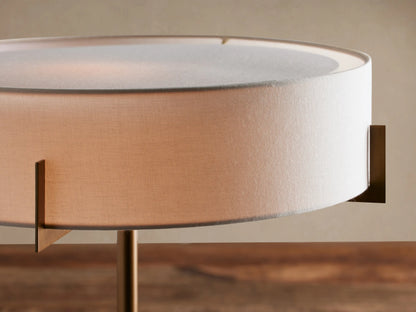Frederic Table Lamp with antiqued brass frame, black granite base, and cylindrical white linen shade, on a wooden table.
