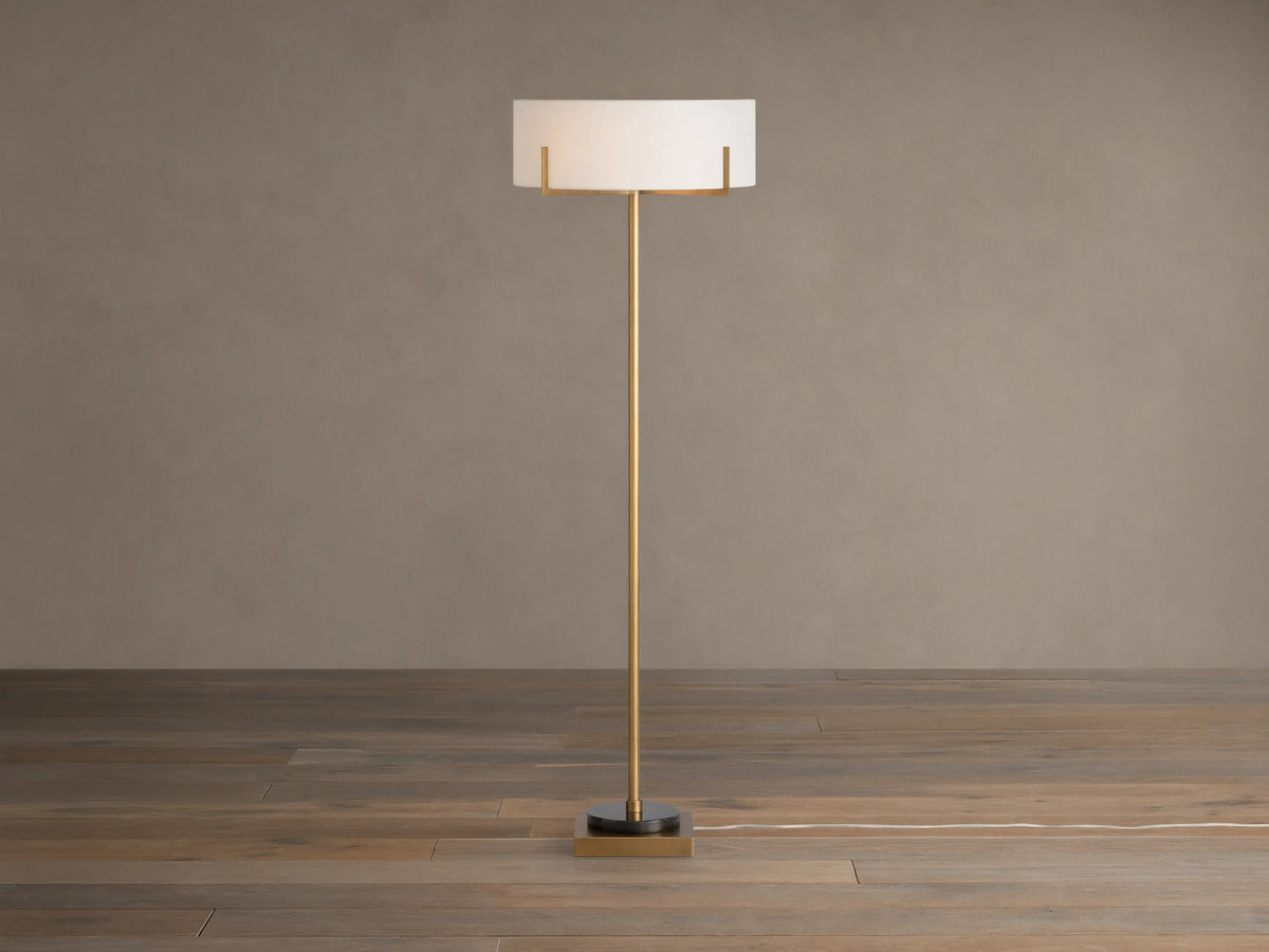 Frederic Floor Lamp: tall antiqued brass pole, white linen shade, black granite base on a wooden floor against a brown wall.
