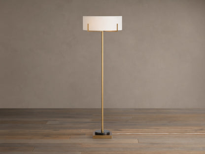 Frederic Floor Lamp: tall antiqued brass pole, white linen shade, black granite base on a wooden floor against a brown wall.