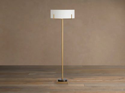 Frederic Floor Lamp: tall antiqued brass pole, white linen shade, black granite base on a wooden floor against a brown wall.