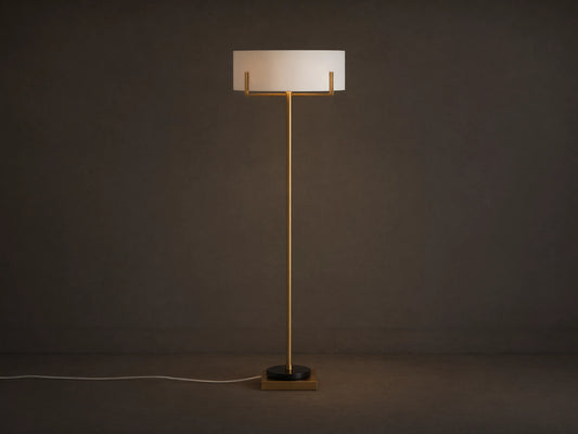 Frederic Floor Lamp: tall antiqued brass pole, white linen shade, black granite base on a wooden floor against a brown wall.