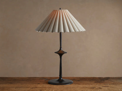 Santiago Table Lamp with a dark bronze base & stem, and a natural pleated linen shade emitting a warm glow, on a wooden table.