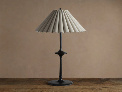 Santiago Table Lamp with a dark bronze base & stem, and a natural pleated linen shade emitting a warm glow, on a wooden table.