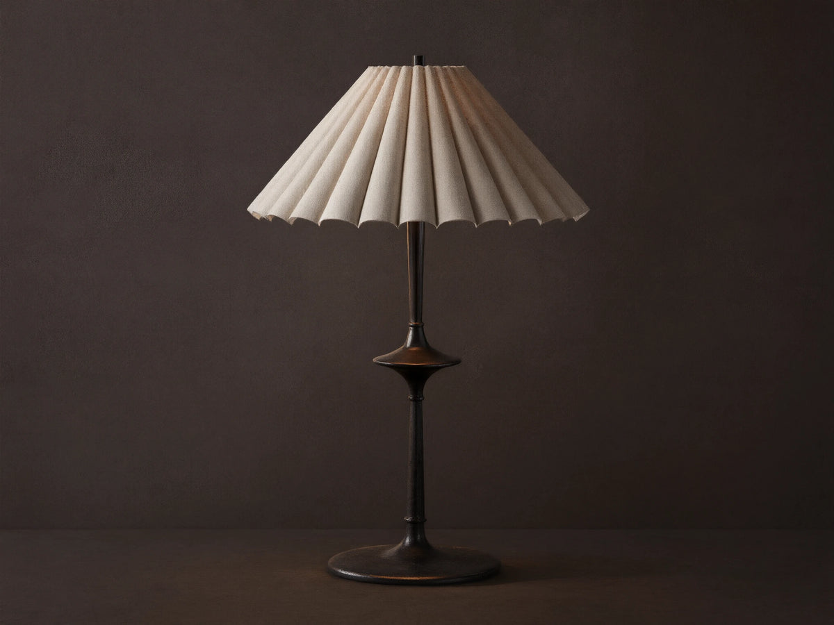 Santiago Table Lamp with a dark bronze base & stem, and a natural pleated linen shade emitting a warm glow, on a wooden table.