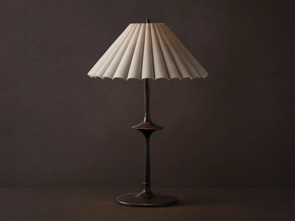 Santiago Table Lamp with a dark bronze base & stem, and a natural pleated linen shade emitting a warm glow, on a wooden table.