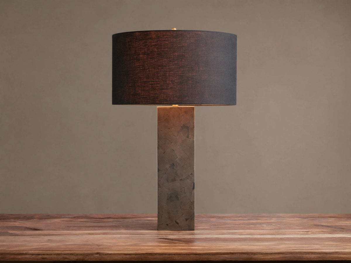 Pyrite Table Lamp with a rectangular base made of dark, shimmering pyrite pieces, topped by a black linen drum shade on a wooden table.