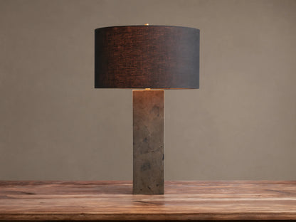 Pyrite Table Lamp with a rectangular base made of dark, shimmering pyrite pieces, topped by a black linen drum shade on a wooden table.
