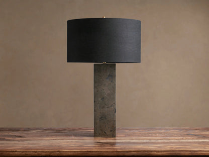 Pyrite Table Lamp with a rectangular base made of dark, shimmering pyrite pieces, topped by a black linen drum shade on a wooden table.