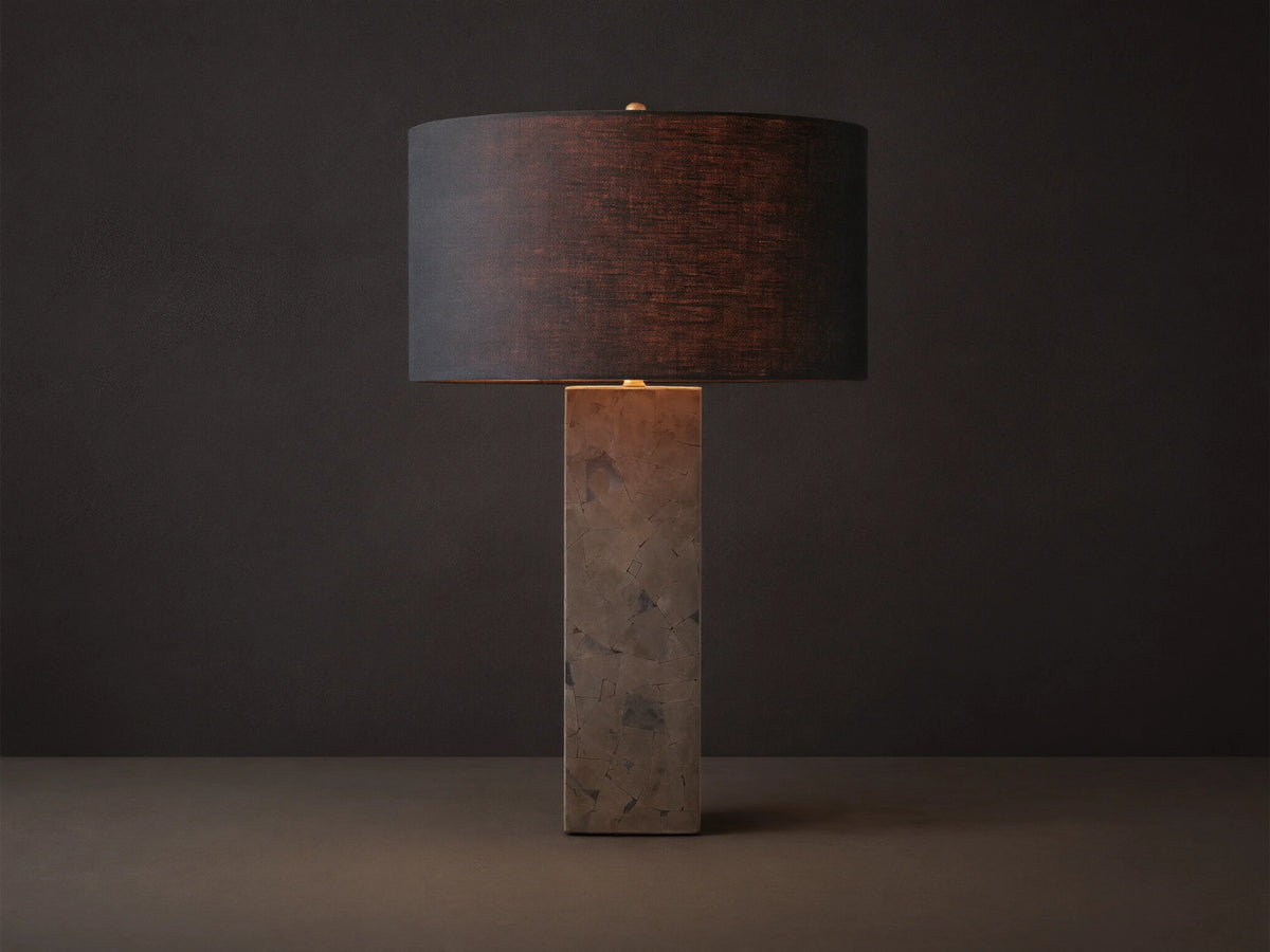 Pyrite Table Lamp with a rectangular base made of dark, shimmering pyrite pieces, topped by a black linen drum shade on a wooden table.