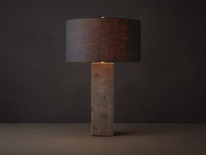 Pyrite Table Lamp with a rectangular base made of dark, shimmering pyrite pieces, topped by a black linen drum shade on a wooden table.