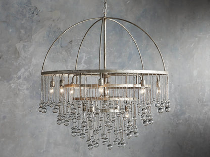 Aubrey Round Chandelier in Silver with a handcrafted iron frame, hanging glass crystals, and five bulbs, set against a grey wall.