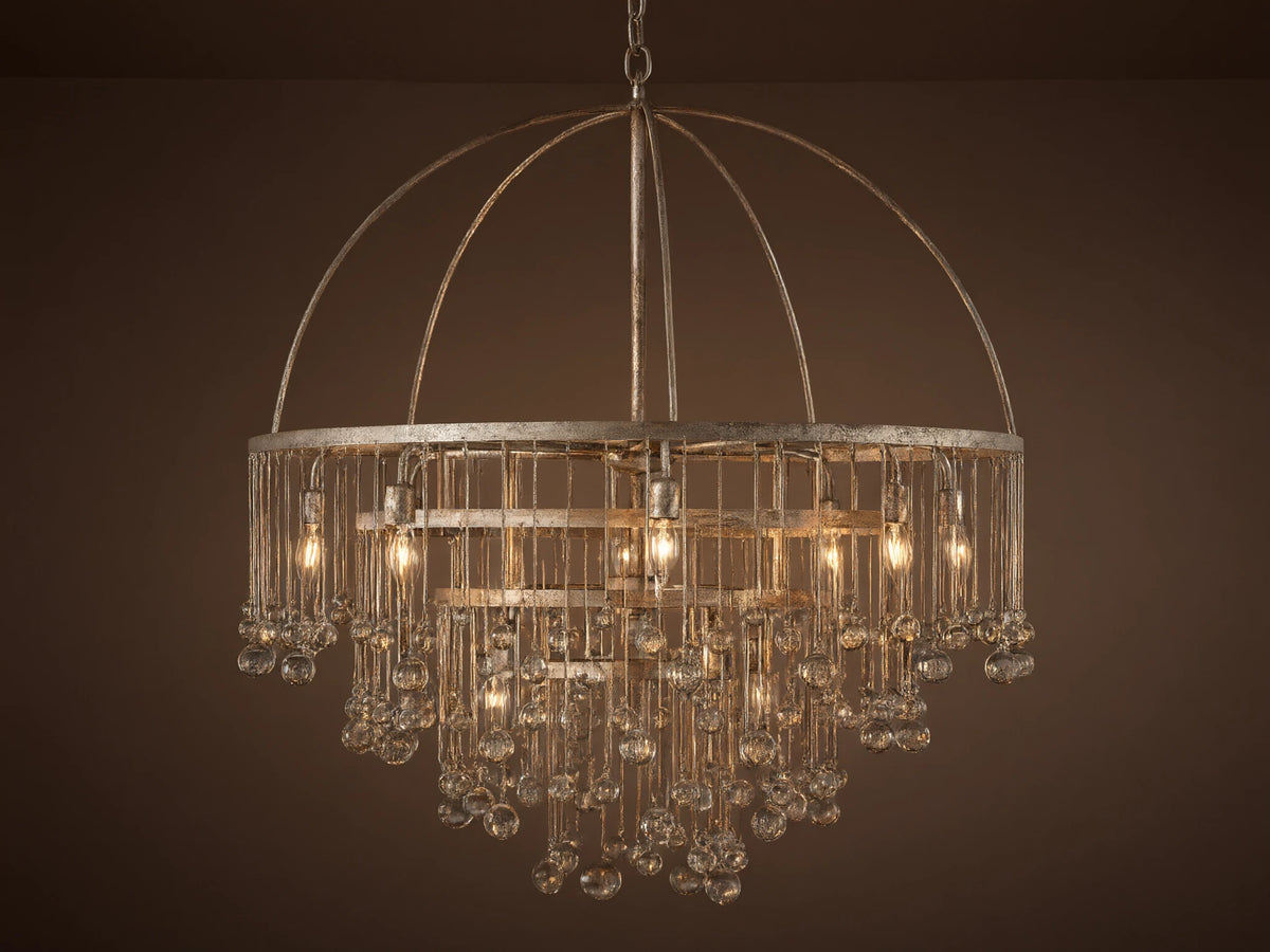 Aubrey Round Chandelier in Silver with a handcrafted iron frame, hanging glass crystals, and five bulbs, set against a grey wall.