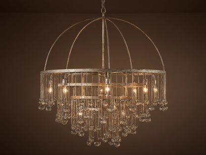 Aubrey Round Chandelier in Silver with a handcrafted iron frame, hanging glass crystals, and five bulbs, set against a grey wall.
