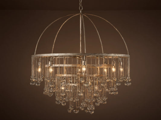 Aubrey Round Chandelier in Silver with a handcrafted iron frame, hanging glass crystals, and five bulbs, set against a grey wall.